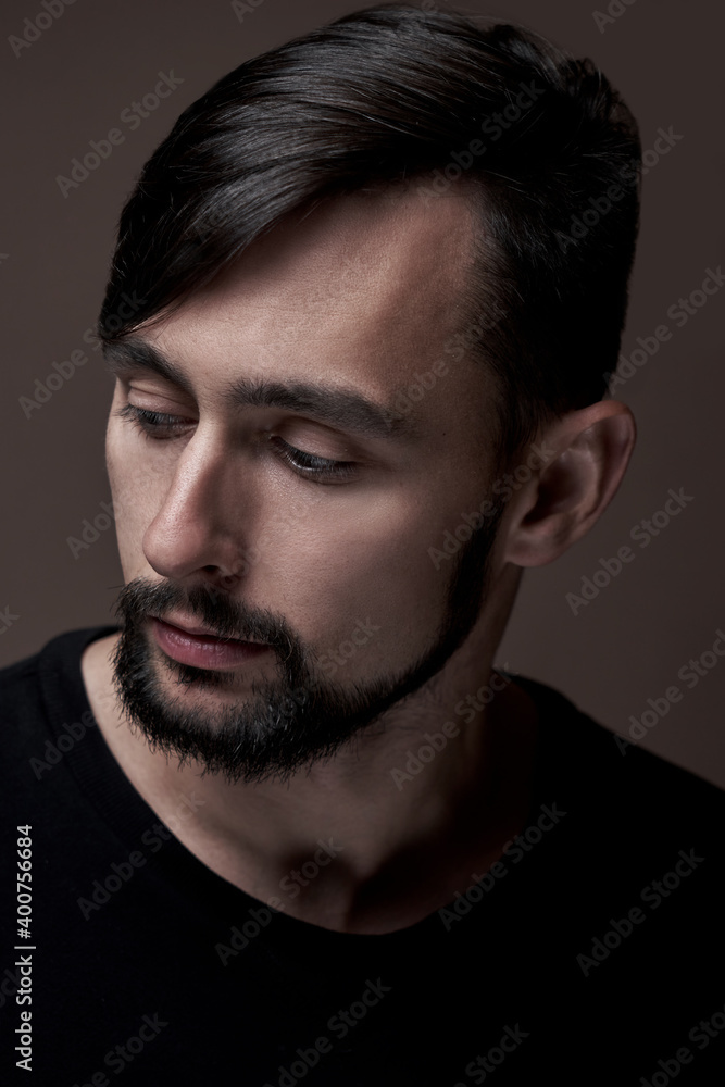 Portrait of handsome caucasian man with beard and stylish haircut in black clothes posing on brown background isolated in studio, fashion model lowering eyes