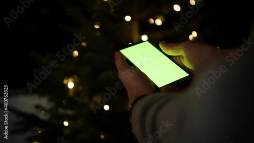 Man Using Smartphone in verticall Mode with Green Mock-up Screen, Doing Swiping, Scrolling Gestures in front of christmas tree. Concept for Application. Gift, Christmas Present. photo
