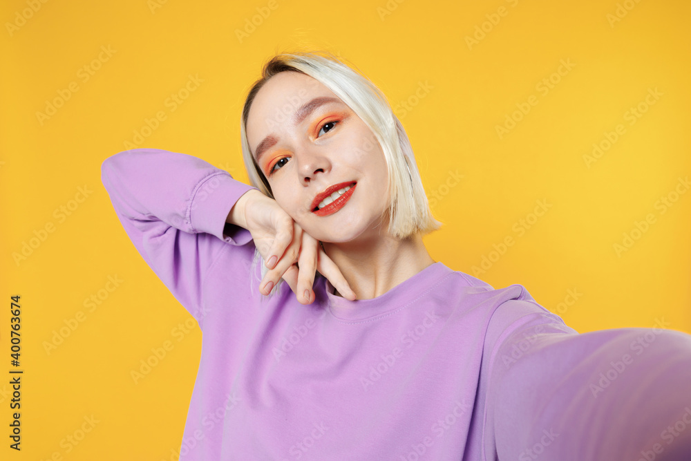 Smiling young blonde caucasian woman 20s bob haircut bright makeup wearing casual basic purple shirt close up doing selfie shot on mobile phone isolated on yellow color background studio portrait. Smiling young blonde caucasian woman 20s bob haircut bright makeup wearing casual basic purple shirt close up doing selfie shot on mobile phone isolated on yellow color background studio portrait.