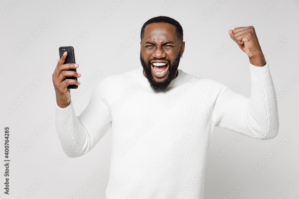 Happy young african american man 20s wearing basic sweater standing using mobile cell phone typing sms message doing winner gesture clenching fists isolated on white color background studio portrait. Happy young african american man 20s wearing basic sweater standing using mobile cell phone typing sms message doing winner gesture clenching fists isolated on white color background studio portrait.