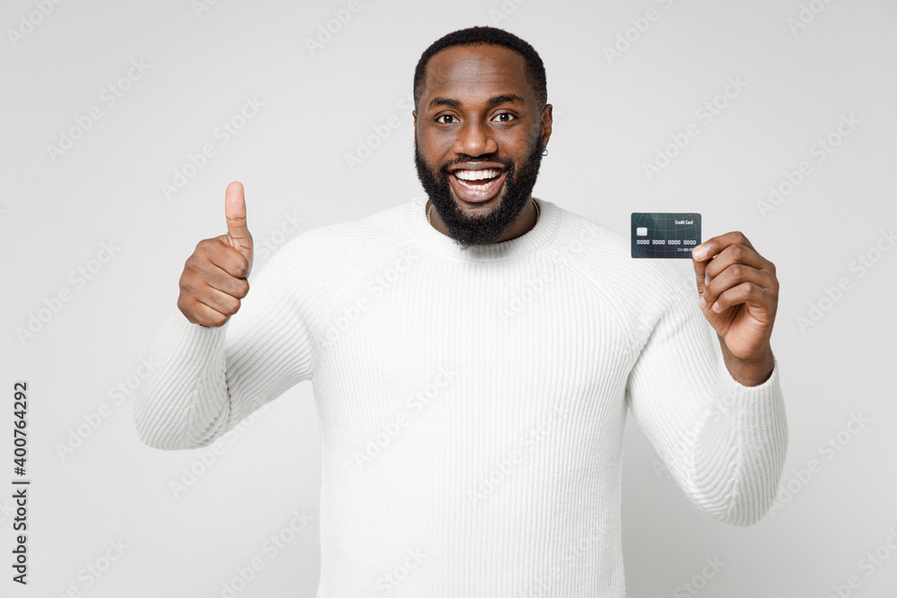 Cheerful smiling young african american man 20s wearing casual basic sweater standing showing thumb up hold credit bank card looking camera isolated on white color wall background studio portrait.