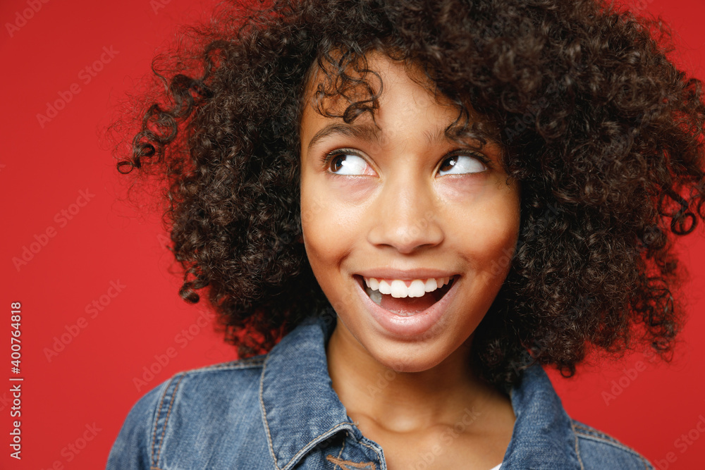 Close up of excited little african american kid girl 12-13 years old in denim jacket keeping mouth open looking aside isolated on red background children studio portrait. Childhood lifestyle concept. Close up of excited little african american kid girl 12-13 years old in denim jacket keeping mouth open looking aside isolated on red background children studio portrait. Childhood lifestyle concept.