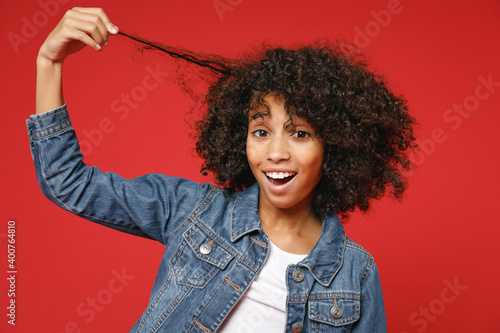 Φωτογραφία Excited funny little african american kid girl 12-13 years old wearing casual denim jacket hold curl hair isolated on bright red color background children studio portrait