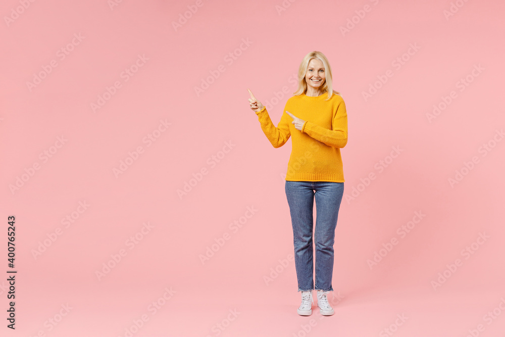 Full length of smiling elderly gray-haired blonde woman lady 40s 50s years old in yellow basic sweater standing point index fingers aside up isolated on pastel pink color background studio portrait. Full length of smiling elderly gray-haired blonde woman lady 40s 50s years old in yellow basic sweater standing point index fingers aside up isolated on pastel pink color background studio portrait.