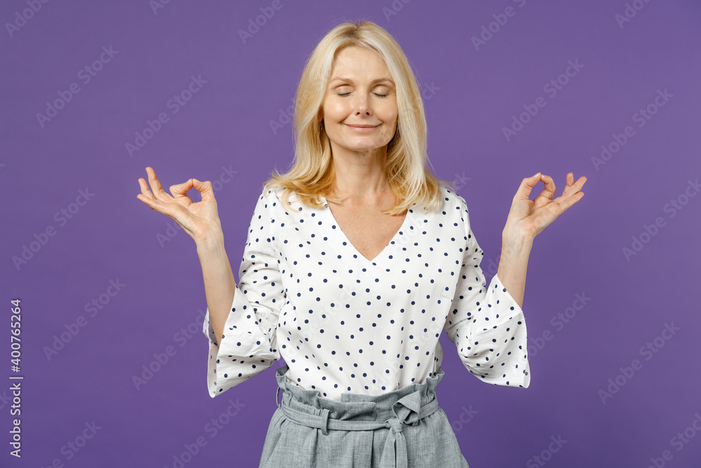 Smiling elderly gray-haired blonde woman lady 40s 50s years old in white dotted blouse hold hands in yoga gesture relaxing meditating trying to calm down isolated on violet background studio portrait. Smiling elderly gray-haired blonde woman lady 40s 50s years old in white dotted blouse hold hands in yoga gesture relaxing meditating trying to calm down isolated on violet background studio portrait.
