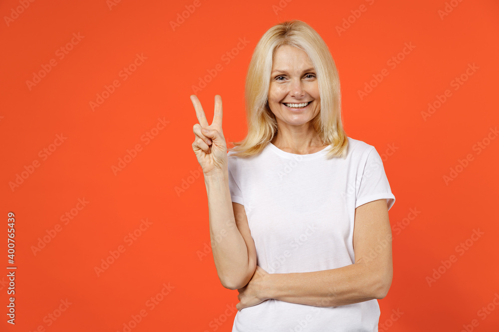 Smiling funny elderly gray-haired blonde woman lady 40s 50s years old in white casual t-shirt standing showing victory sign looking camera isolated on bright orange color background studio portrait. - obrazy, fototapety, plakaty 