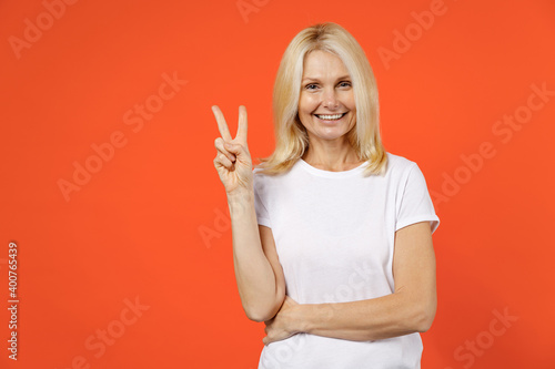 Smiling funny elderly gray-haired blonde woman lady 40s 50s years old in white casual t-shirt standing showing victory sign looking camera isolated on bright orange color background studio portrait.