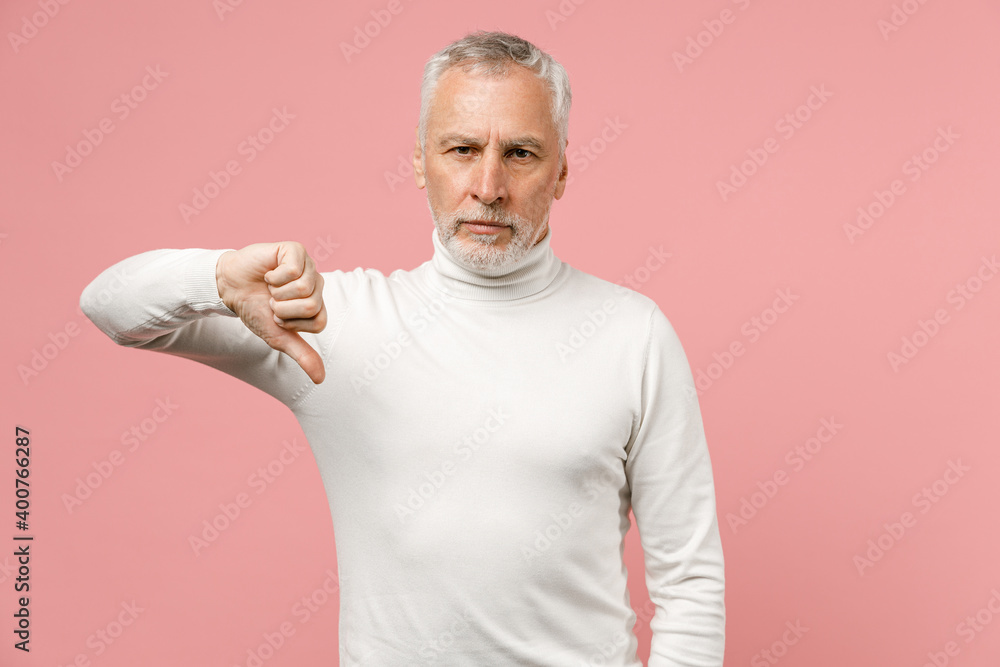 Dissatisfied displeased elderly gray-haired mustache bearded man in casual basic white turtleneck standing showing thumb down looking camera isolated on pastel pink color background studio portrait. Dissatisfied displeased elderly gray-haired mustache bearded man in casual basic white turtleneck standing showing thumb down looking camera isolated on pastel pink color background studio portrait.