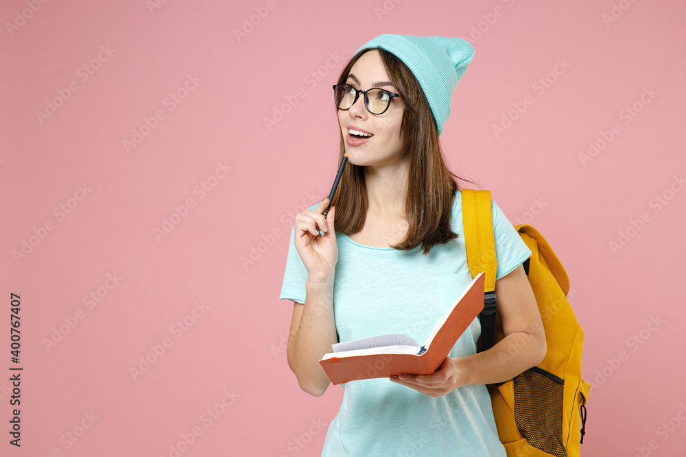Pensive young woman student in casual blue t-shirt hat glasses backpack writing notes in notebooks isolated on pastel pink color background studio. Education in high school university college concept. Pensive young woman student in casual blue t-shirt hat glasses backpack writing notes in notebooks isolated on pastel pink color background studio. Education in high school university college concept.