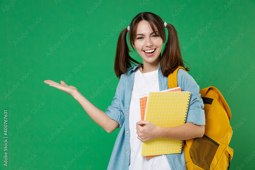 Cheerful funny young woman student in casual blue shirt backpack hold notebooks pointing hand aside isolated on green background studio portrait. Education in high school university college concept. Cheerful funny young woman student in casual blue shirt backpack hold notebooks pointing hand aside isolated on green background studio portrait. Education in high school university college concept.