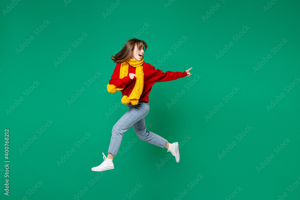 Full length side view excited young brunette woman 20s in knitted red sweater yellow scarf jumping like running pointing index finger aside isolated on bright green color background studio portrait.
