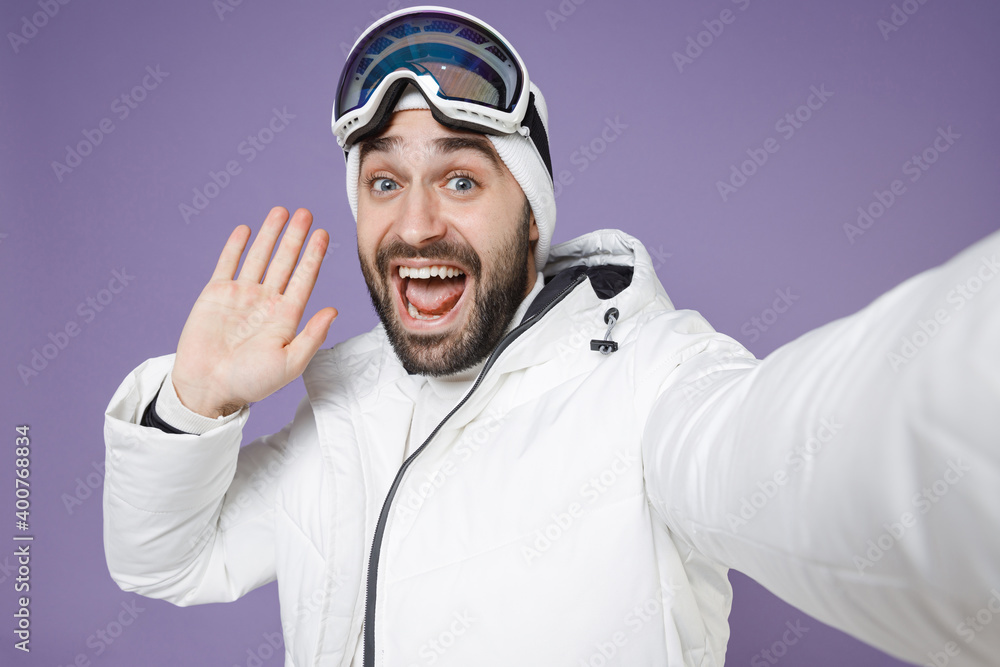Close up of amazed skier man in windbreaker jacket ski goggles mask doing selfie shot on mobile phone greeting with hand spend weekend in mountains isolated on purple background. People hobby concept. Close up of amazed skier man in windbreaker jacket ski goggles mask doing selfie shot on mobile phone greeting with hand spend weekend in mountains isolated on purple background. People hobby concept.