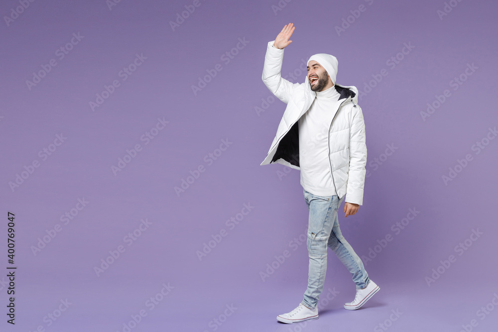 Full length side view funny man in white windbreaker jacket hat waving greeting with hand as notices someone isolated on purple background studio portrait. People lifestyle cold winter season concept. Full length side view funny man in white windbreaker jacket hat waving greeting with hand as notices someone isolated on purple background studio portrait. People lifestyle cold winter season concept.