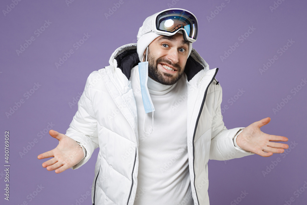 Confused skier man in warm white windbreaker jacket ski goggles face mask safe from coronavirus virus covid-19 spreading hands weekend in mountains isolated on purple background. People hobby concept. Confused skier man in warm white windbreaker jacket ski goggles face mask safe from coronavirus virus covid-19 spreading hands weekend in mountains isolated on purple background. People hobby concept.