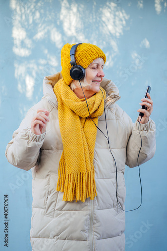 Senior female listening to music and using smartphone. Optimistic elderly woman in trendy outerwear smiling while listening to music and browsing smartphone against blue wall