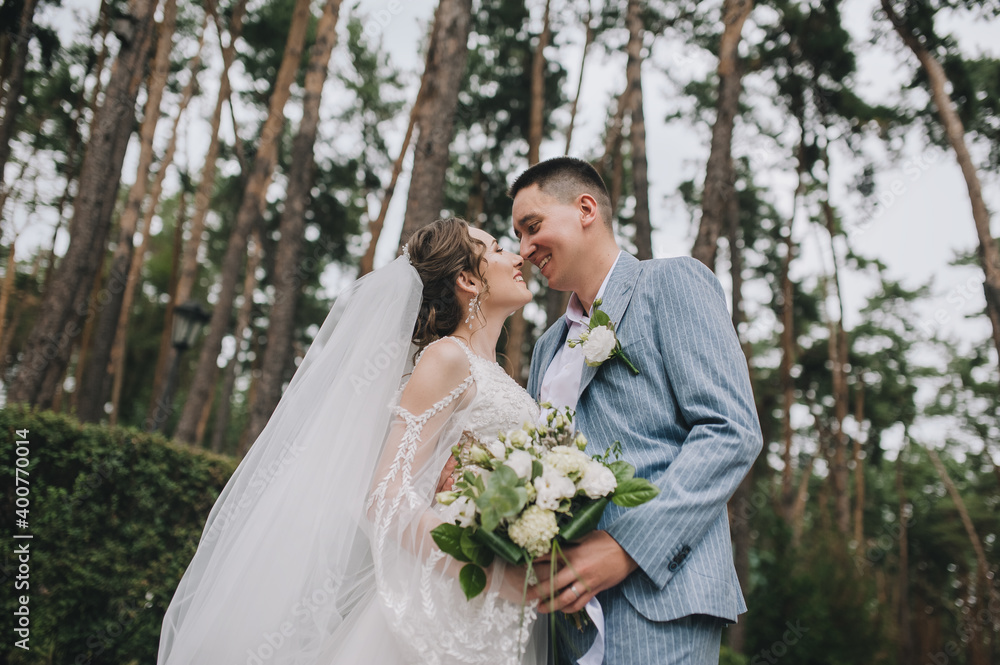 Stylish groom in a blue suit and a beautiful bride in a white lace dress tenderly hug and smile in the forest, in nature with tall pines. Wedding portrait of the newlyweds. Stylish groom in a blue suit and a beautiful bride in a white lace dress tenderly hug and smile in the forest, in nature with tall pines. Wedding portrait of the newlyweds.