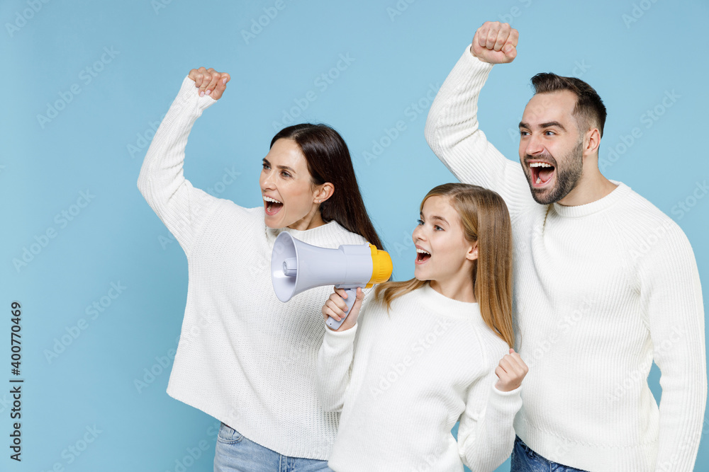 Excited young parents mom dad with child kid daughter teen girl in sweaters screaming in megaphone looking aside clenching fists isolated on blue background. Family day parenthood childhood concept.