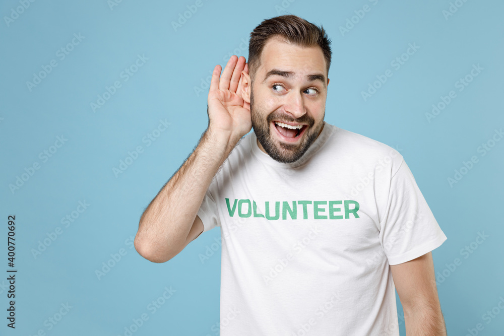 Excited curious young man in white volunteer t-shirt try to hear you overhear listening intently isolated on blue background studio portrait. Voluntary free work assistance help charity grace concept. Excited curious young man in white volunteer t-shirt try to hear you overhear listening intently isolated on blue background studio portrait. Voluntary free work assistance help charity grace concept.