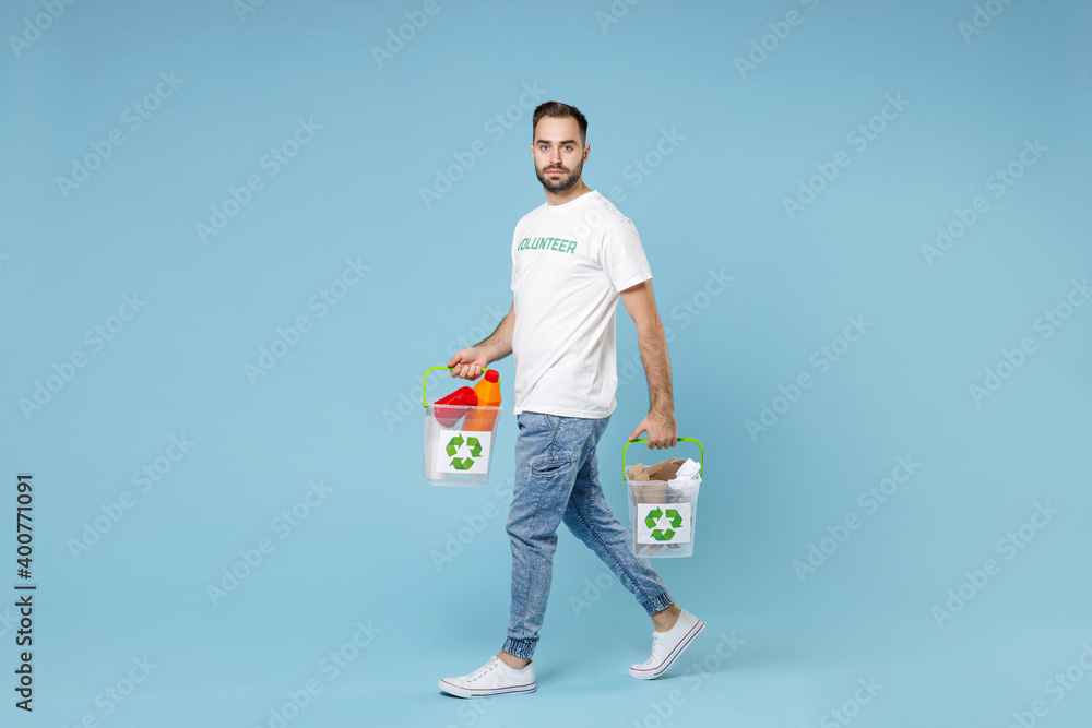 Full length of serious young man in volunteer t-shirt hold baskets with plastic paper trash isolated on blue background studio portrait. Voluntary free assistance help trash sorting recycling concept. Full length of serious young man in volunteer t-shirt hold baskets with plastic paper trash isolated on blue background studio portrait. Voluntary free assistance help trash sorting recycling concept.