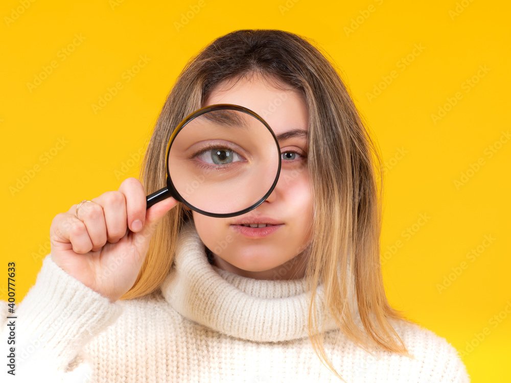 Girl holds a magnifying glass in front of her eye and looks at camera. Observant young woman with magnifying glass. Exploring what's around us. Detailed review of interesting object. Girl holds a magnifying glass in front of her eye and looks at camera. Observant young woman with magnifying glass. Exploring what's around us. Detailed review of interesting object.