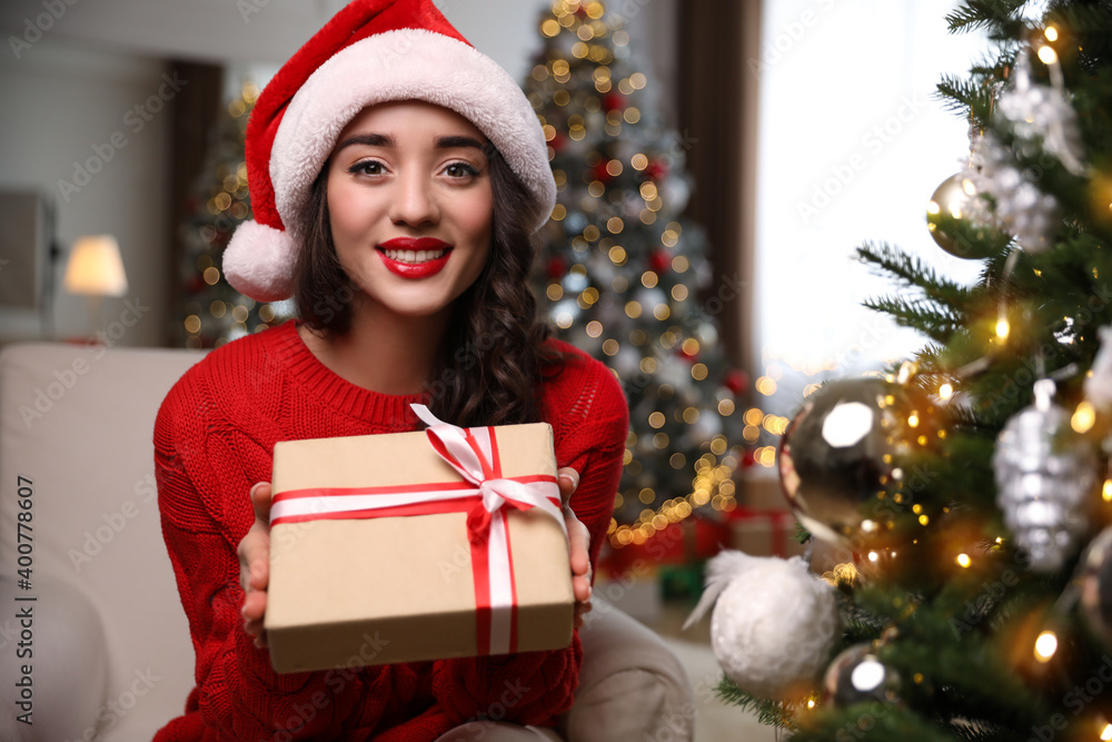 Beautiful woman in Santa hat with gift box near Christmas tree at home