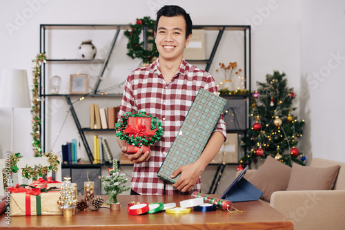 Happy handsome young Asian man standing at table with wrapped presents and Christmas wreath in hands