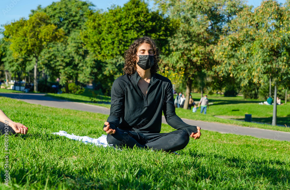 Fototapeta premium Young Attractive Man with Mask Meditating in a Yoga Class at the Park in Summer