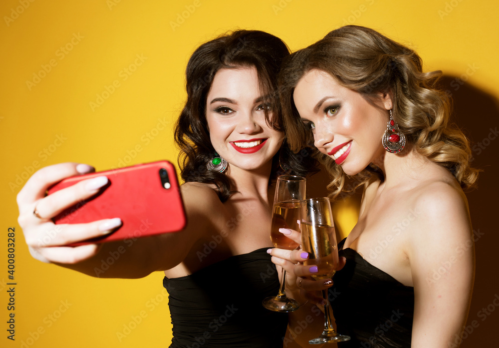 Celebration, party, and people concept - two chic young women in black cocktail dresses drinking champagne and taking a selfie.