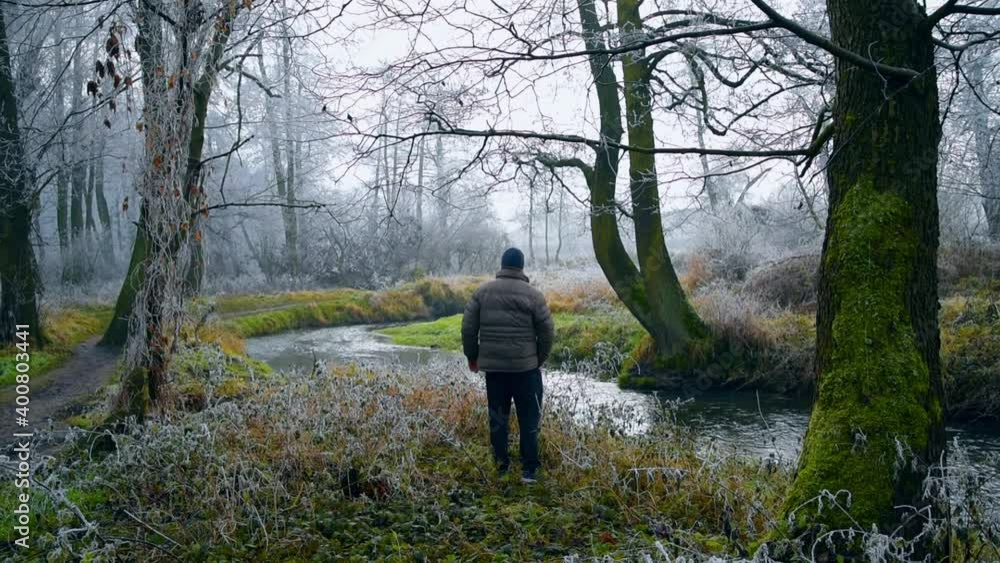 Man dressed in a thick jacket walking through the park among trees and frosted plants on a gray cloudy day. A man resting in the park among green, frozen grasses and branches. Early winter landscape. Man dressed in a thick jacket walking through the park among trees and frosted plants on a gray cloudy day. A man resting in the park among green, frozen grasses and branches. Early winter landscape.