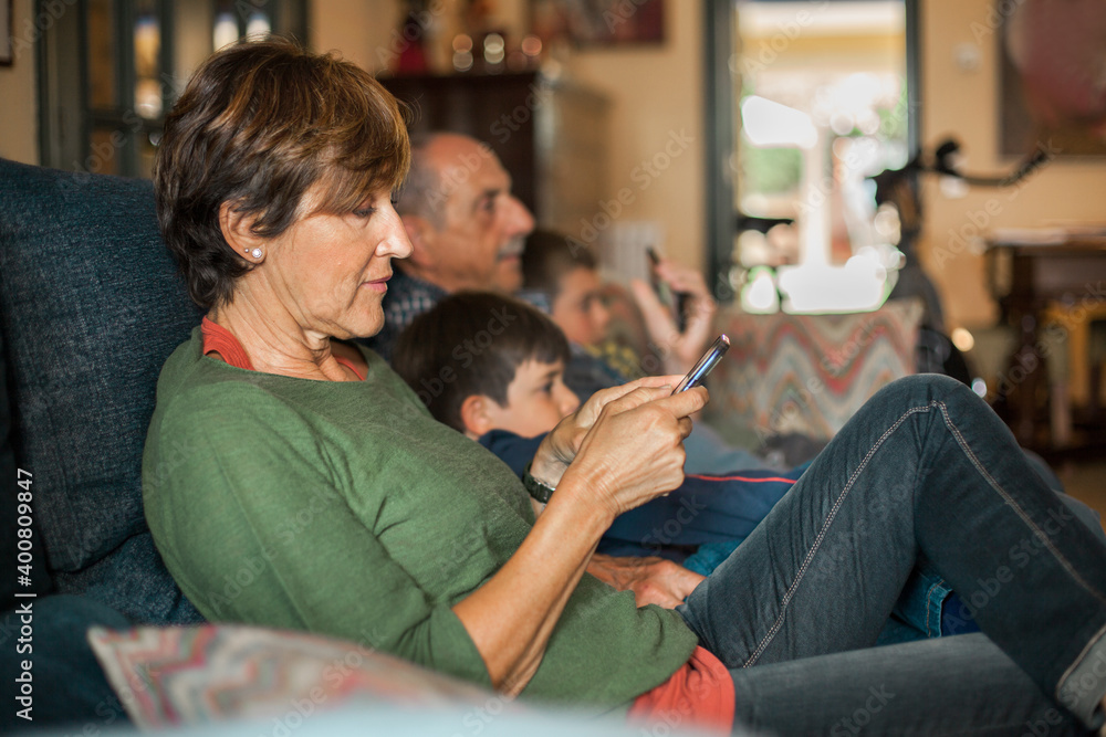 Grandmother sitting on the sofa takes a look at her cell phone while the family watches television. Grandmother sitting on the sofa takes a look at her cell phone while the family watches television.
