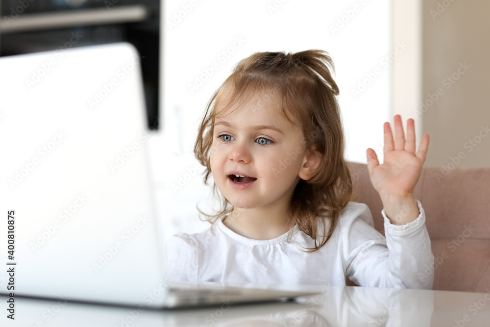 A little smiling adorible girl uses a laptop video chat for communicion while sitting at a table at home. Child online. A little smiling adorible girl uses a laptop video chat for communicion while sitting at a table at home. Child online.