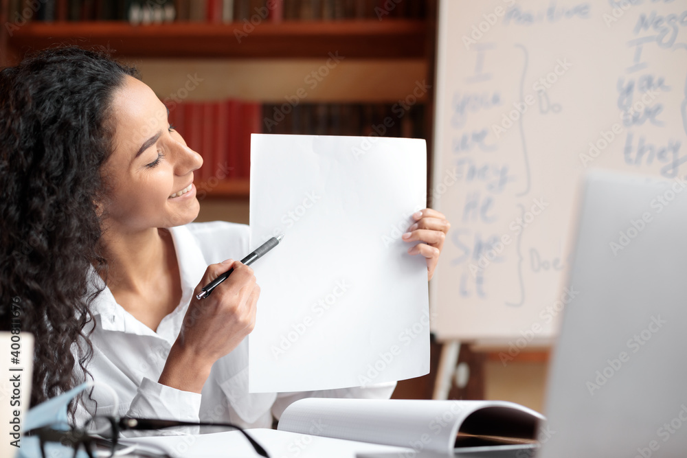 Woman sitting at desk, having videocall on laptop, holding paper Woman sitting at desk, having videocall on laptop, holding paper