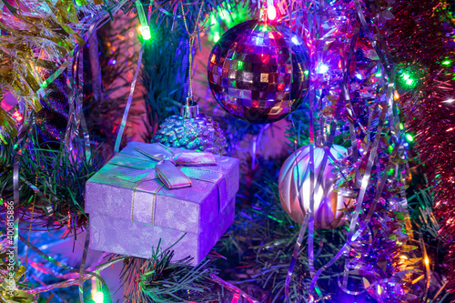 Gift box with a bow on the branches of the Christmas tree illuminated with colorful lights garlands.