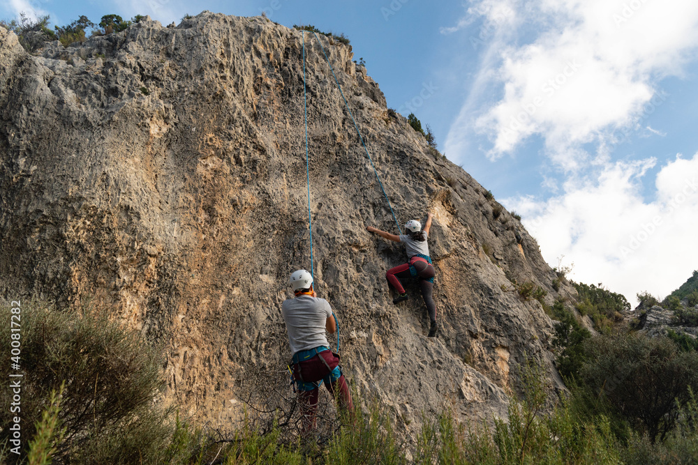 Bottom perspective of a girl climber and a boy belaying her as she climbs a stone wall outdoors