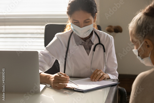Young female GP or therapist in medical facial mask consult mature patient about health insurance in hospital. Woman doctor in facemask consider results or treatment with elderly client in clinic.