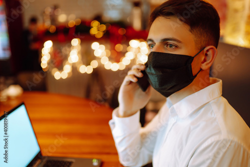 Young man wearing medical face mask working on laptop computer and speaks on the phone at home during winter holiday. The concept of preventing the spread of the epidemic coronavirus. Remote work. 
