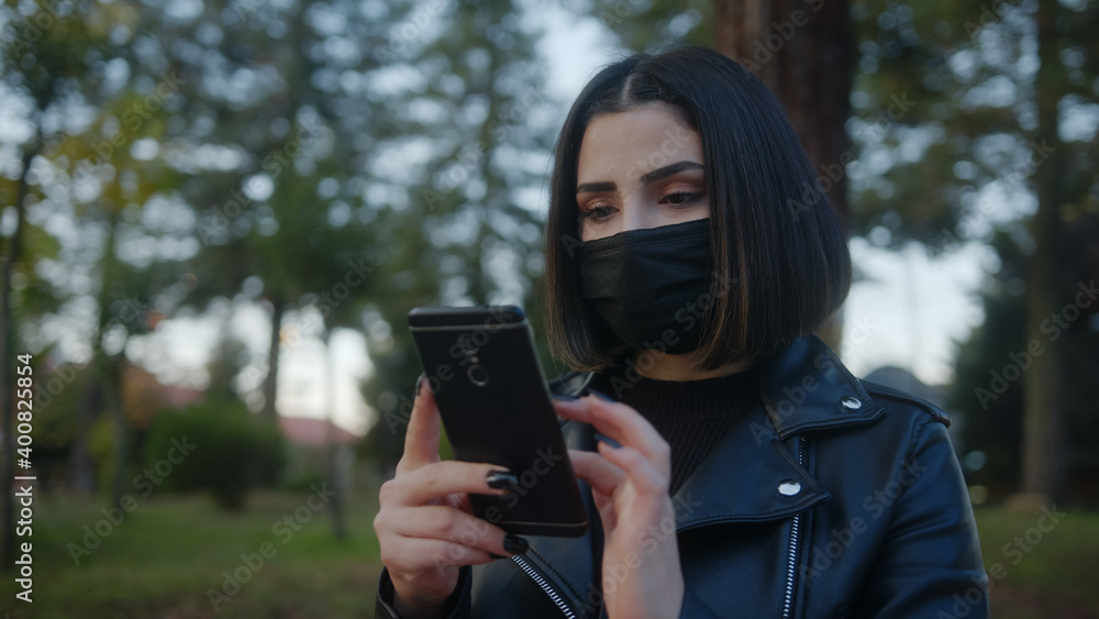 With leather coat beautiful young woman black wear face mask and use of mobile phone in park. Covid-19 Concept.