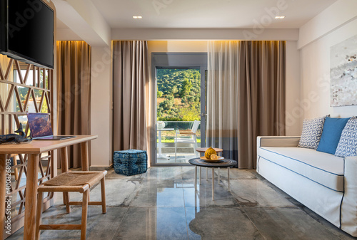 Modern interior of contemporary living room with handmade wicker wood furniture, beige blue soft textile, grey marble tile floor, summer garden view in open French window terrace