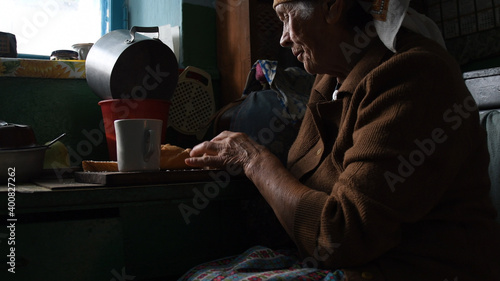 Old wrinkled woman tilts her head and takes bread by hand. Suffering loneliness at senior age. Portrait of rural elderly woman in contour sunlight. Concept of poverty and geriatric health problem