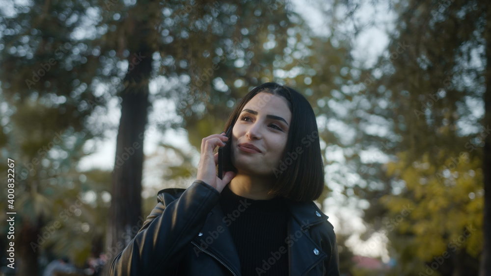 Among green trees young woman walking in the park, talking nicely on the phone and smiles, enjoys a conversation with her beloved or friend. Relations at a distance, making phone call.