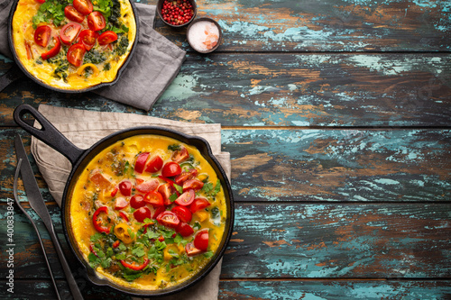 Healthy frittata in two cast iron pans with fried beaten eggs and seasonal vegetables on rustic wooden background. Italian omelette with organic spinach, bell pepper, tomatoes, top view 