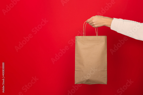 Female hand hold large gift bag made of brown craft paper on red background.