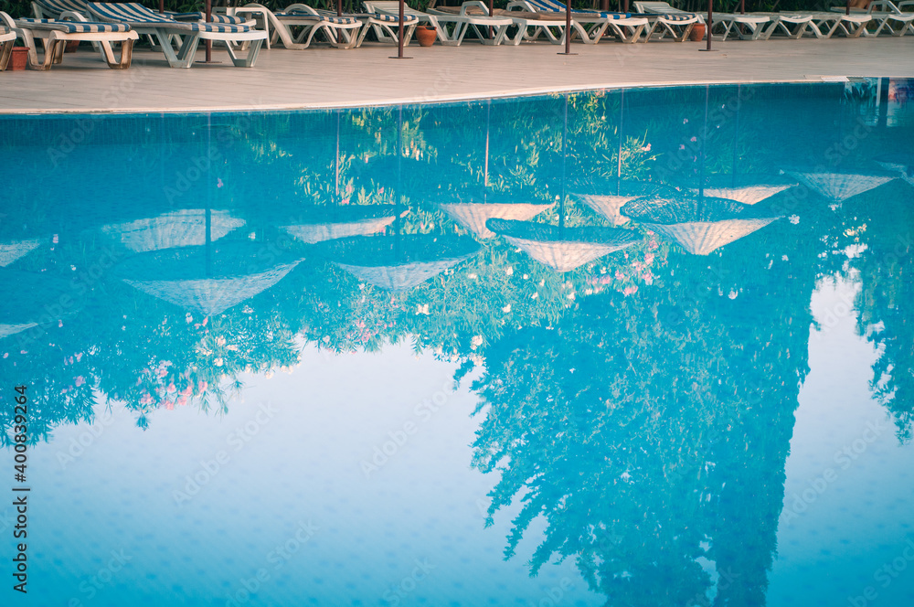 Transparent blue pool with sun loungers in the early morning at the resort at the hotel. Reflection in umbrellas and greenery in calm water.