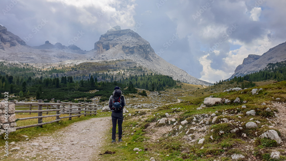 A man with a big hiking backpack hiking along a gravelled road in Italian Dolomites. There is a wooden fence along the road. Sharp and stony mountain chains in front. Few boulders on the green meadow.