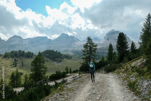 A woman with a hiking backpack hiking on a gravelled road in high Italian Dolomites. There is a dence forest at the foothill, and steep and sharp mountain chain in the back. Discovering and exploring