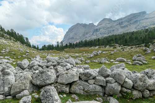 A stony wall made of bigger lose boulders on a lush green meadow in Italian Dolomites. There are sharp and stony mountains in the back. The mountains are surrounded by clouds. Remote and desolate area © Chris