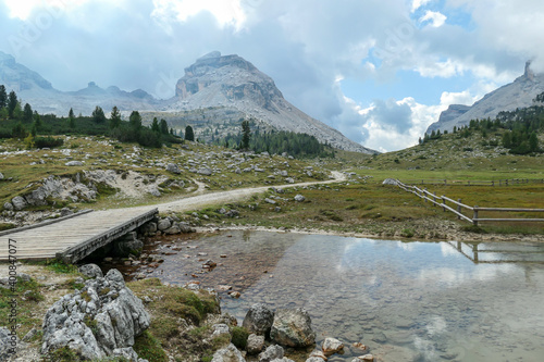 A small pond under a wooden bridge in Italian Dolomites. The pond is reflecting the surrounding mountains and the sky. High mountain chains in the back. Forest on the side. Desolated and remote area