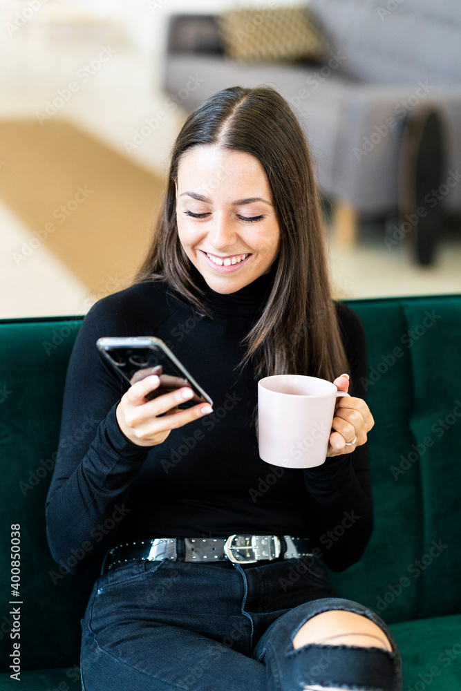 Smiling young female influencer holding smart phone and coffee cup while sitting on sofa at loft apartment Smiling young female influencer holding smart phone and coffee cup while sitting on sofa at loft apartment