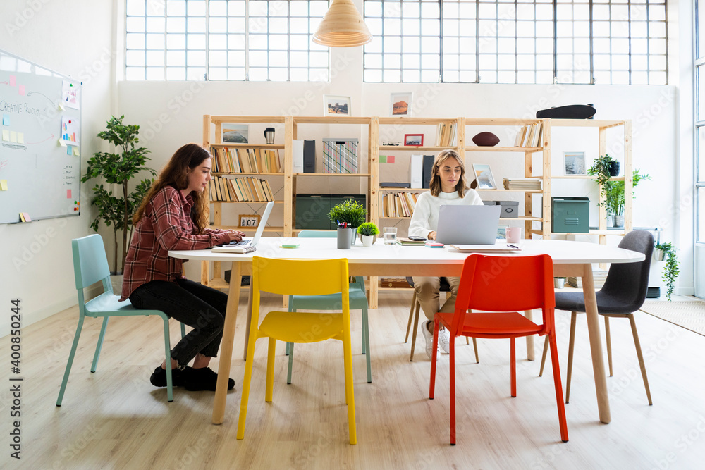 Businesswoman and colleague working on laptop while sitting by table at office Businesswoman and colleague working on laptop while sitting by table at office