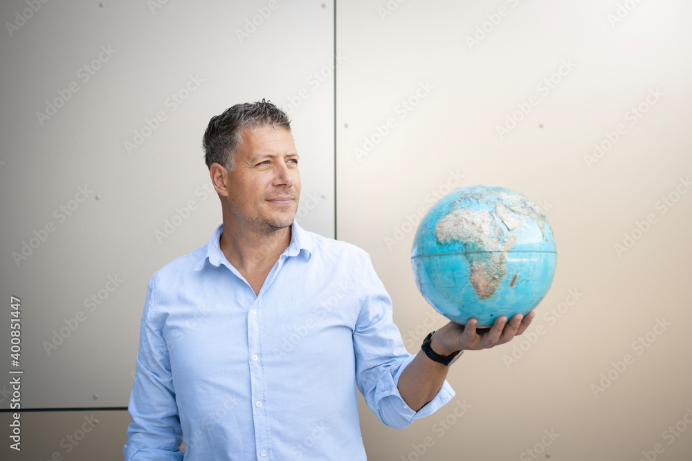 Businessman looking away while holding globe standing against wall Businessman looking away while holding globe standing against wall